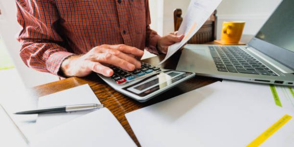 Wide angle image depicting a senior man's hand holding an energy bill while the other hand checks the numbers on a calculator. The table is strewn with documents and also a laptop. Room for copy space.